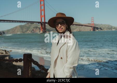 Vue latérale d'une jeune femme souriante dans une tenue tendance avec chapeau et lunettes de soleil debout sur une rive contre le Golden Gate Bridge en Californie par beau temps Banque D'Images