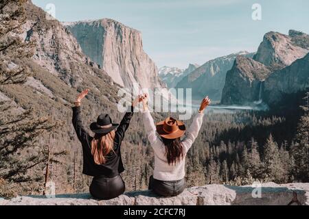 Vue arrière de amies méconnues dans des vêtements tendance et des chapeaux assis sur le bord de la falaise rocheuse et en appréciant Paysages de montagne étonnants lors de la visite du parc national de Yosemite en Californie en plein soleil le printemps Banque D'Images