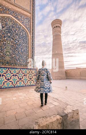 Vue arrière de la femelle en manteau ornemental debout sur une place âgée contre ciel nuageux à Boukhara, Ouzbékistan Banque D'Images