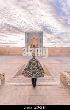 Vue arrière de la femelle en manteau ornemental debout sur une place âgée contre ciel nuageux à Boukhara, Ouzbékistan Banque D'Images