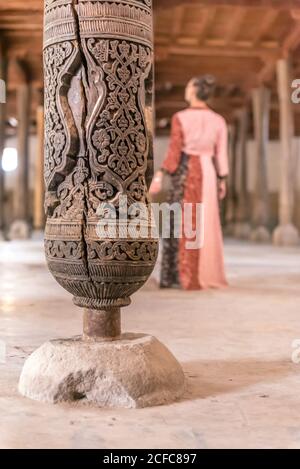 Colonne en bois avec décoration orientale sculptée majestueuse dans la mosquée Juma avec une femme floue dans des vêtements traditionnels debout et regardant loin Banque D'Images