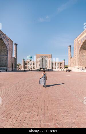 Vue arrière de la femme anonyme en robe marchant sur la place pavée près des bâtiments islamiques traditionnels tout en visitant le Registan à Samarkand, Ouzbékistan Banque D'Images