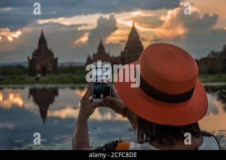 Vue arrière d'une femme en chapeau orange se tenant au bord du lac et prenant des photos avec un smartphone des beaux temples bouddhistes de Bagan Birmanie avec ciel nuageux au coucher du soleil en arrière-plan Banque D'Images