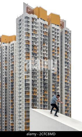 Homme décontracté sans visage dans des vêtements décontractés prétendant surfer sur toit en pente de béton contre l'extérieur des gratte-ciel résidentiels avec gris Et des taches jaunes à Hong Kong Banque D'Images