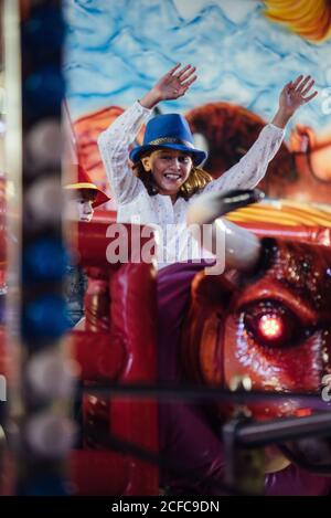 Bonne fille aux bras relevés souriant et à cheval mécanique de taureau tout en s'amusant dans un parc d'attractions Banque D'Images