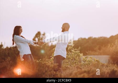 Vue latérale d'un couple heureux amoureux assorti à des vêtements décontractés tenir les mains et regarder l'un l'autre en se tenant debout champ herbacé au coucher du soleil Banque D'Images