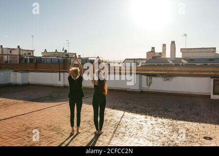 Vue arrière du corps entier d'une femme mûre avec une jeune fille en vêtements de sport noirs debout en position de salut vers le haut avec les bras relevés tout en faisant la séquence de salutation du soleil pendant l'entraînement de yoga sur le toit Banque D'Images
