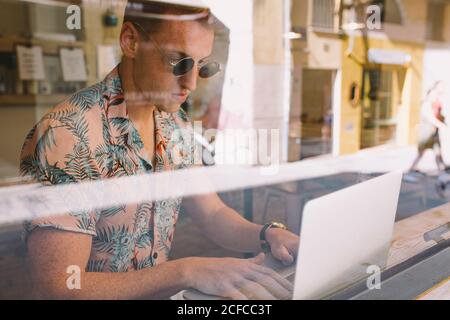 Jeune homme concentré travaillant sur un ordinateur portable à table tout au long d'un fenêtres dans le café Banque D'Images