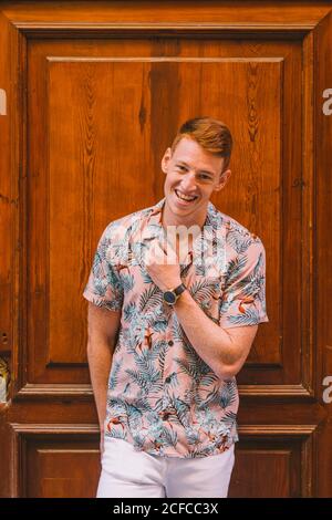 Beau homme heureux homme dans une chemise hawaïenne penchée sur du bois porte regardant l'appareil photo sourire Banque D'Images