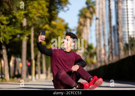Joyeux jeune homme ethnique dans les vêtements de sport souriant et prenant le selfie en vous asseyant sur la piste pendant votre entraînement physique par beau temps en ville Banque D'Images