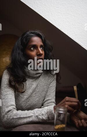 Vue latérale de la jeune femme indienne calme en robe décontractée reposant sur un canapé confortable près de la fenêtre dans la chambre moderne mansardée tout en roulant de la marijuana cigaret Banque D'Images