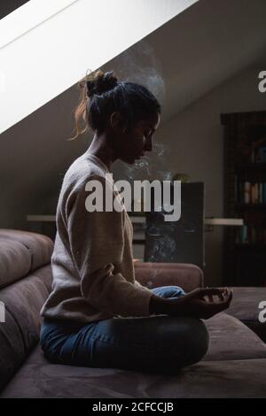 Vue latérale de la jeune femme indienne assise avec les yeux fermés dans lotus poser sur le canapé et faire du yoga tout en appréciant odeur agréable de bâton d'encens Banque D'Images