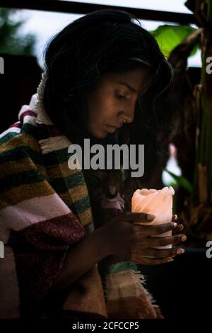 Shaman féminin ethnique jeune concentré dans un poncho chaud tenant une bougie tout en exécutant la cérémonie spirituelle en soirée Banque D'Images