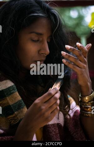 Spirituelle jeune femme ethnique avec les yeux fermés dans les vêtements traditionnels tenir l'encens de sauge brûlant avec de la fumée et méditer à l'extérieur Banque D'Images