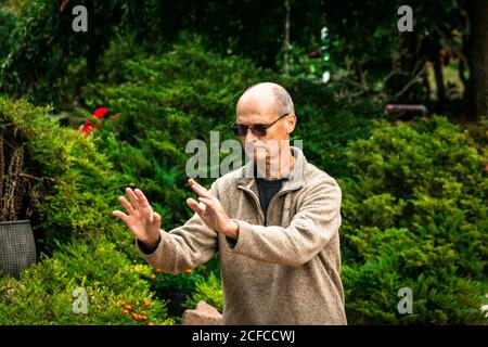 Chauve senior mâle dans des lunettes de soleil exécutant l'exercice pendant les arts martiaux entraînement sur fond flou d'arbres verts dans le jardin Banque D'Images