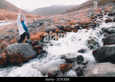Jeune femme aux mains levées méditant et debout sur des rochers près d'une rivière étroite entre les collines Banque D'Images