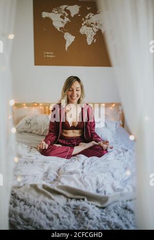 Belle jeune femme en vêtements de sport gardant les yeux fermés et méditant assis dans lotus poser sur un lit confortable le matin Banque D'Images