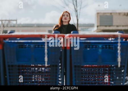 Belle jeune femme attentionnés avec des cheveux rouges et beaucoup de chariots dans le parking par temps nuageux Banque D'Images