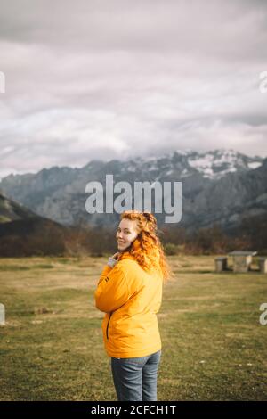 Vue arrière d'une femme souriante dans une veste jaune chaude regardant l'appareil photo sur l'épaule dans un pré d'herbe verte entouré de montagnes enneigées Banque D'Images