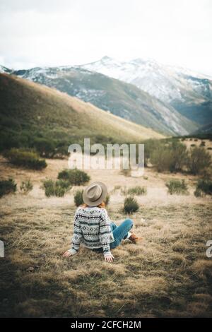 Vue arrière d'une charmante femme pensive méconnaissable avec des cheveux rouges dans un chandail chaud assis sur une pelouse en montagne par temps gris Banque D'Images