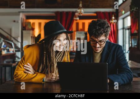 Homme intelligent dans des lunettes et femme élégante dans un chapeau regardant un moniteur pendant que l'homme tape sur un ordinateur portable dans un café Banque D'Images