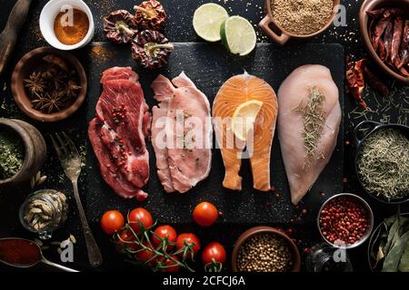 Vue de dessus du filet de bœuf et de porc avec poulet et poisson sur planche à découper en bois noir en composition avec différentes épices aromatiques et assaisonnements sur la table Banque D'Images