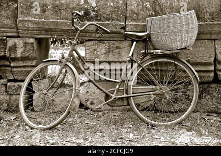 Une image en noir et blanc d'un vieux vélo rustique en fer avec un panier tissé en rotin, appuyé contre un vieux mur de pierre recouvert de lichen. Banque D'Images
