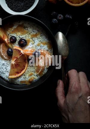 De dessus chia graines pudding avec des tranches d'orange, mandarines et olives dans le bol près de la main saisir une cuillère et pamplemousse sur table sombre Banque D'Images