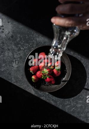 Du dessus de la main de récolte de la bouteille de retenue humaine et verser liquide de fraise fraîche dans un bol illuminé par des rayons de soleil obscurité Banque D'Images