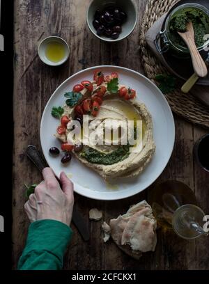 De dessus plaque de délicieux pesto hoummus décoré avec de la cerise tomates et haricots avec persil placés sur un plan d'examen en bois près pain plat et huile Banque D'Images