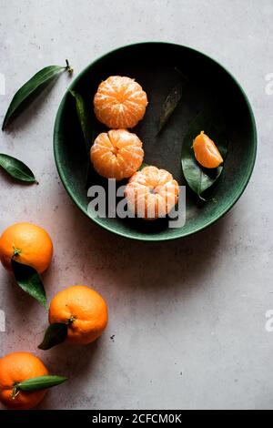 Vue de dessus d'un bol en céramique verte avec des mandarines fraîchement pelées placé sur une table blanche près des fruits non pelés avec des feuilles vertes Banque D'Images