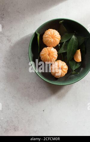 Vue de dessus d'un bol en céramique verte avec des mandarines fraîchement pelées placé sur une table blanche près des fruits non pelés avec des feuilles vertes Banque D'Images