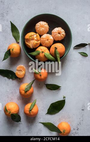 Vue de dessus d'un bol en céramique verte avec des mandarines fraîchement pelées placé sur une table blanche près des fruits non pelés avec des feuilles vertes Banque D'Images