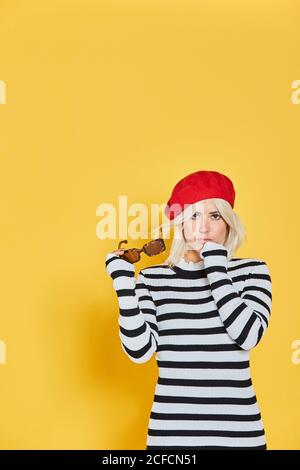 Femme blonde gaie en chemisier rayé et casquette française rouge souriant sur fond jaune Banque D'Images