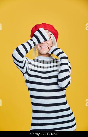 Blonde gaie femme en chemisier rayé et casquette française rouge souriant avec les mains sur le visage sur fond jaune Banque D'Images