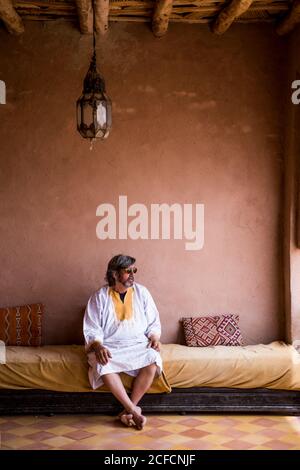 Homme adulte en longs vêtements assis sur un canapé sur la terrasse avec clôture en pierre de style oriental, Maroc Banque D'Images