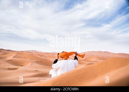 Vue arrière de l'homme portant une longue tenue ajustant un turban coloré assis sur des dunes de désert de sable fin, Maroc Banque D'Images