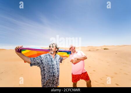 Heureux surpoids mature hommes ayant du plaisir avec le drapeau LGBT pendant sur fond de désert aride et de ciel bleu Banque D'Images