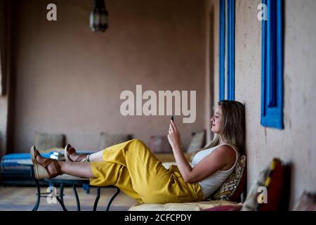 Femme adulte assise sur un canapé en terrasse de style oriental et utilisant un téléphone portable au Maroc Banque D'Images