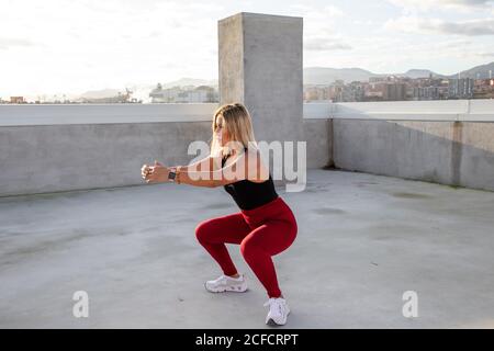 Femme aux cheveux blonds concentrée en tenue active en position de squat avec bras étirés sur le toit du bâtiment Banque D'Images