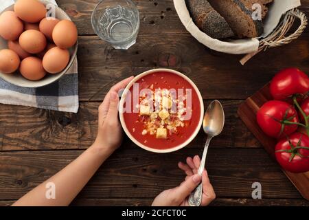 D'en haut de la photo d'une personne méconnaissable tenant un bol de gazpacho rouge dans une table rustique avec des arrangements d'oeufs, de pain, et de tomates fraîches Banque D'Images