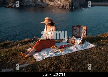 Vue latérale de la femme en chapeau avec livre assis sur le tapis pour pique-nique avec pastèque et verre de boisson sur la côte sèche et romantique le long de l'eau sereine Banque D'Images