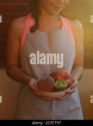 Des pêches appétissantes savoureuses et des kiwis juteux mûrs coupés dans un bol entre les mains d'une femme souriante en tablier Banque D'Images