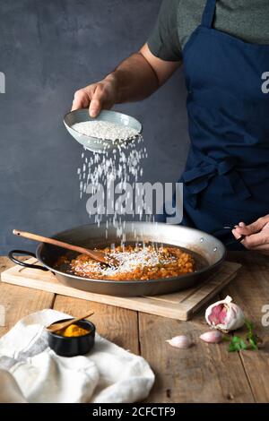 Crop man dans le tablier bleu foncé ajoutant le riz blanc à ingrédients grillés hachés sur une grande casserole en métal tout en préparant du délicieux plat savoureux à la maison Banque D'Images