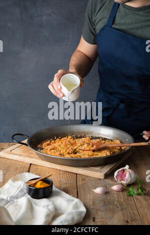 Crop man dans le tablier bleu foncé versant l'huile au riz avec des ingrédients grillés hachés sur une grande casserole en métal pendant la cuisson plat savoureux appétissant à la maison Banque D'Images