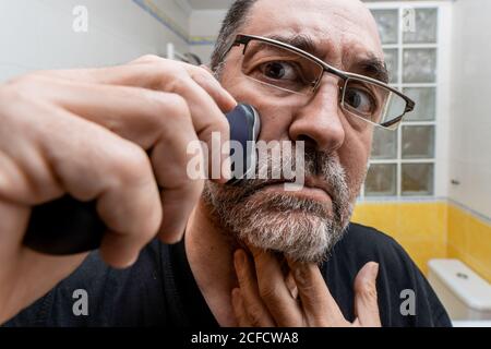 Gros plan d'un homme à barbe d'âge moyen et sérieusement concentré dans des lunettes rasage avec rasoir électrique devant le miroir dans la salle de bains Banque D'Images