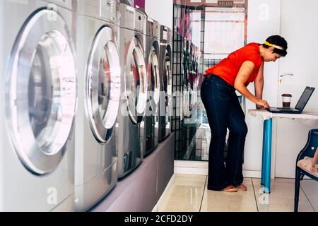 Jeune mode mode brunette utiliser un ordinateur portable et attendre dans un magasin de service de tapis de blanchisserie dans la ville - lavage activité vêtements dans la boutique automatique - Banque D'Images