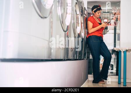 Jeune mode tendance brunette utiliser le téléphone et attendre dans un magasin de service de buanderie dans la ville - lavage des vêtements activité dans la boutique automatique - moderne Banque D'Images