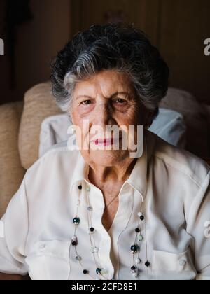 Portrait d'une femme heureuse senior à cheveux gris et boucles en chemise blanche et avec des perles sur le cou en regardant l'appareil photo à la maison Banque D'Images