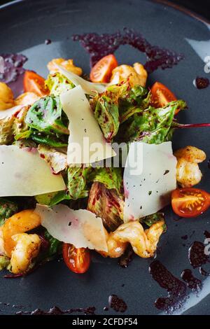 De dessus gros plan de salade délicieuse avec des légumes frais et feuilles avec crevettes et fromage servies sur une assiette noire avec sauce Banque D'Images